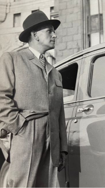 Archival black-and-white portrait of a man in a suit and hat beside a car.