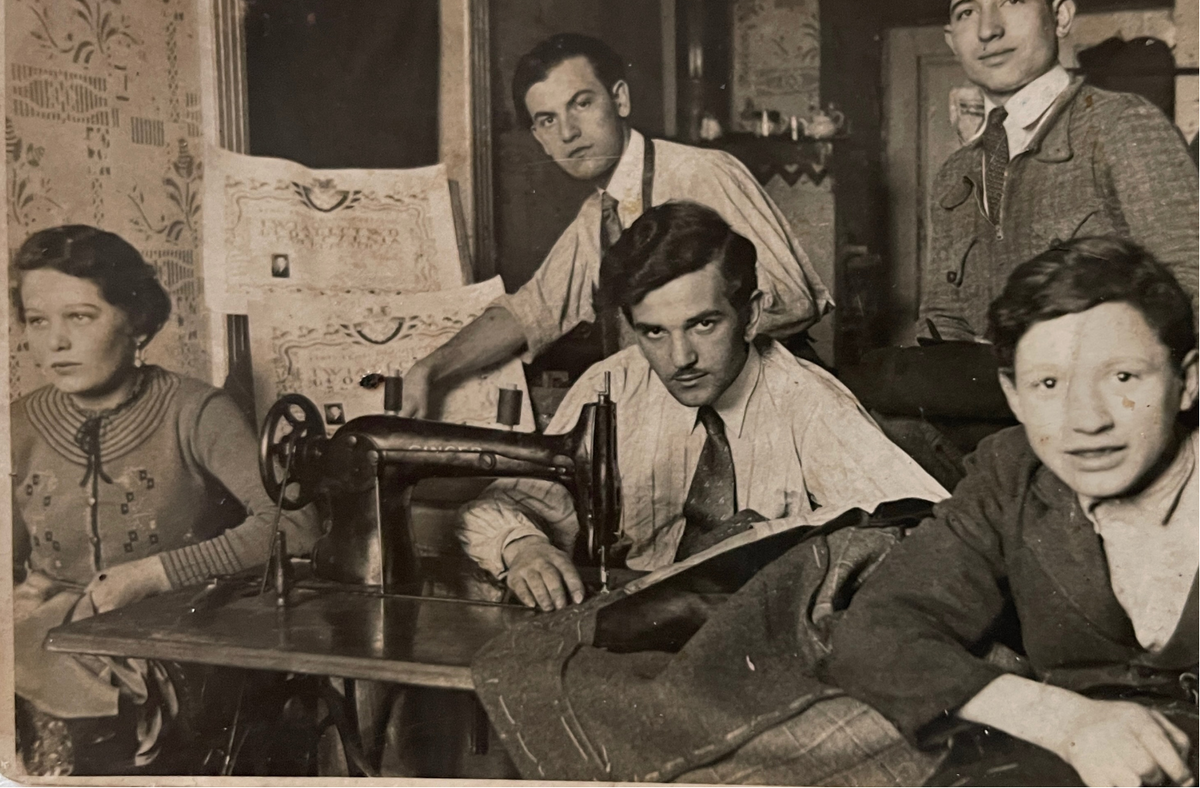 Archival photograph of a tailoring workshop with a group gathered around a sewing machine.