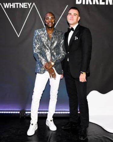 Two guests at a Whitney event, one in a metallic patterned jacket and white trousers, the other in a black tuxedo.